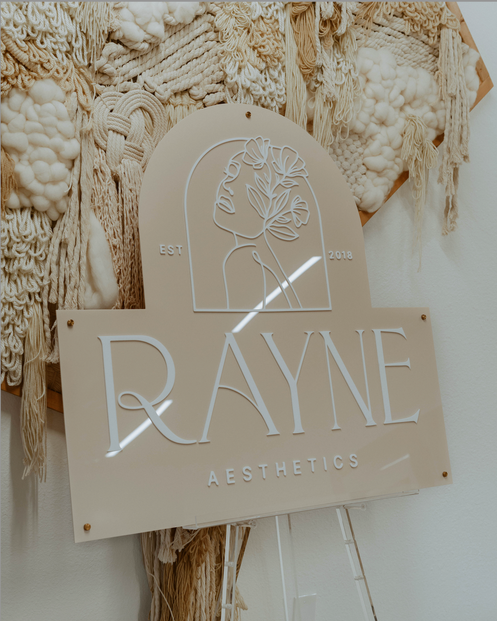 Nude Acrylic Logo Sign with text that reads Rayne Aesthetics featuring a hand-drawn woman's face with flower line art. 